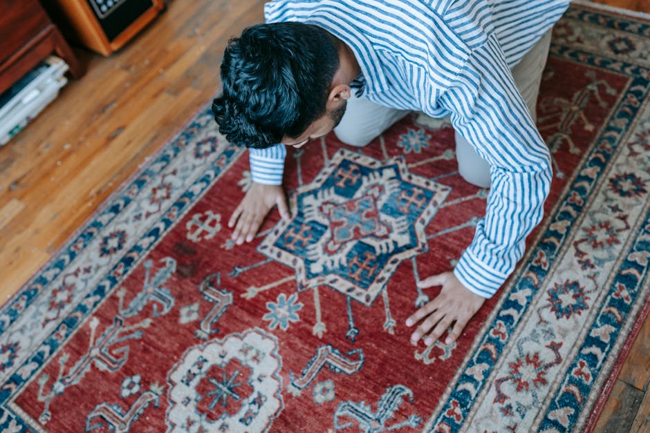 A woman with brown hair tied back, dressed in a light blue and white striped shirt and blue jeans, is kneeling on a patterned rug in a residential bedroom. She is using a blue cloth to clean or dust t