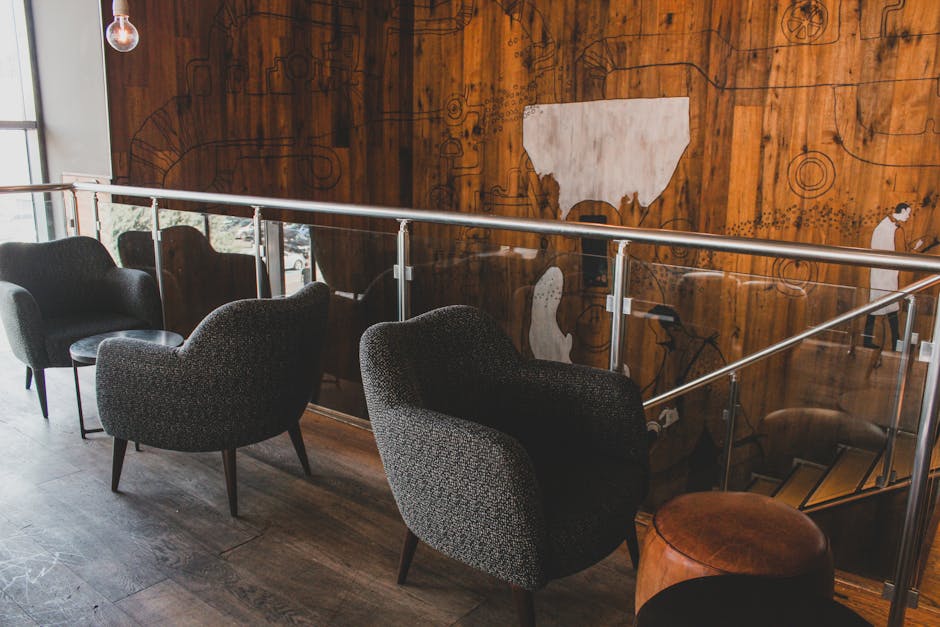 A row of four dark upholstered armchairs with tapered wooden legs arranged along a glass safety railing in a modern interior space. The chairs are placed on a wooden floor with visible grain, and ther