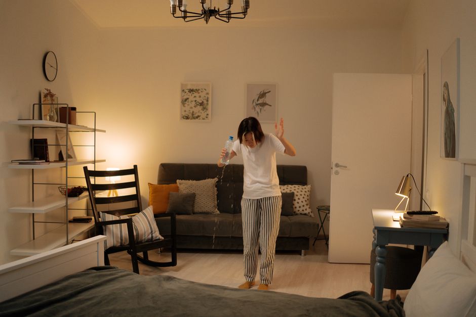 A woman in striped pajama bottoms and a white t-shirt stands in a modern living room, captured in the process of cleaning with a spray bottle. She appears to be addressing a spill on the dark gray sof