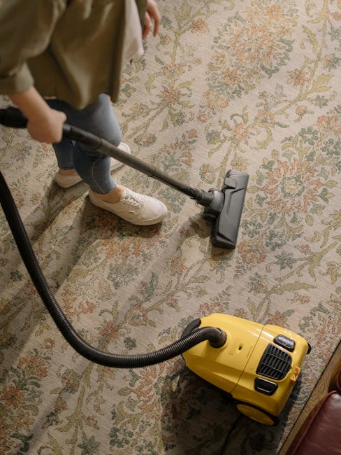 A woman in striped pajama bottoms and a white t-shirt stands in a modern living room, captured in the process of cleaning with a spray bottle. She appears to be addressing a spill on the dark gray sof